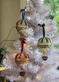 Three decorative hot air balloon ornaments hanging on a white artificial Christmas tree with warm string lights. The ornaments feature intricate beadwork and embroidery in blue, red, green, and gold tones.