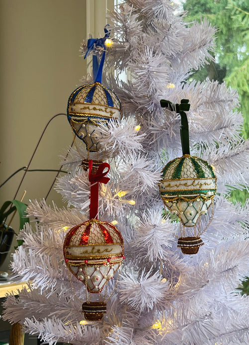 Three decorative hot air balloon ornaments hanging on a white artificial Christmas tree with warm string lights. The ornaments feature intricate beadwork and embroidery in blue, red, green, and gold tones.
